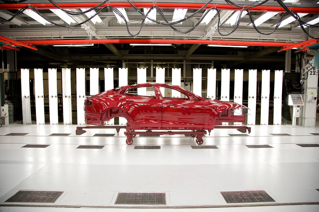 The outer part of a red car is in an assembly line