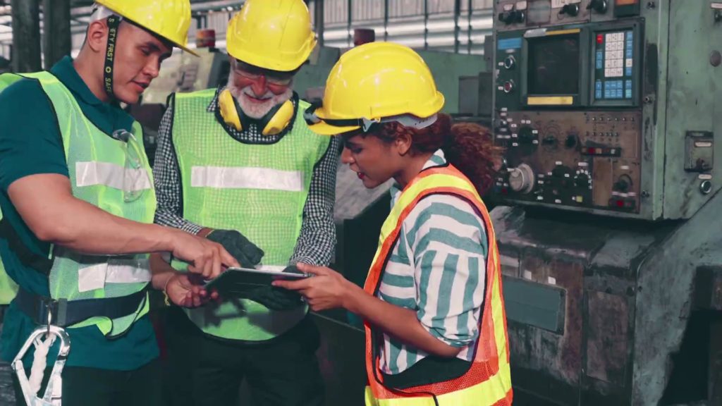 Two male and one female factory workers are looking at an black ipad discussing the quality assurance process in manufacturing.